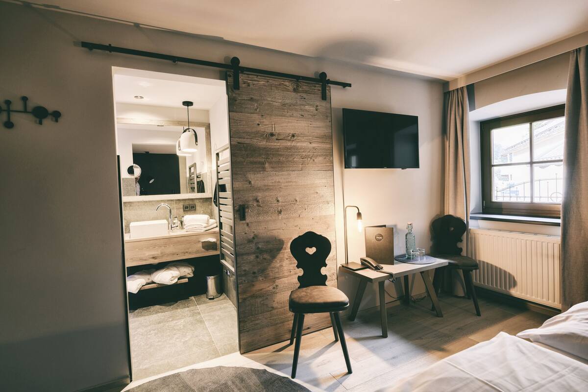 A hotel room with a wooden sliding door leading to a bathroom, a desk with a chair, a TV mounted on the wall, and a window with curtains.