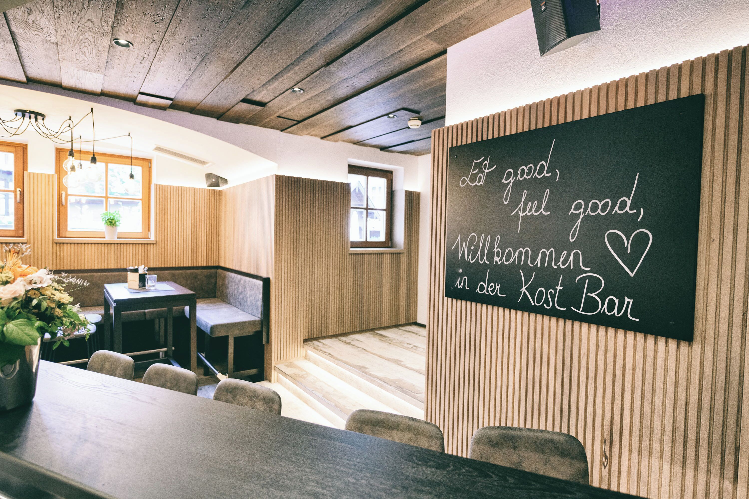 Een restaurantinterieur met houten lambrisering, een schoolbord met een tekst en een zithoek met tafels en stoelen.