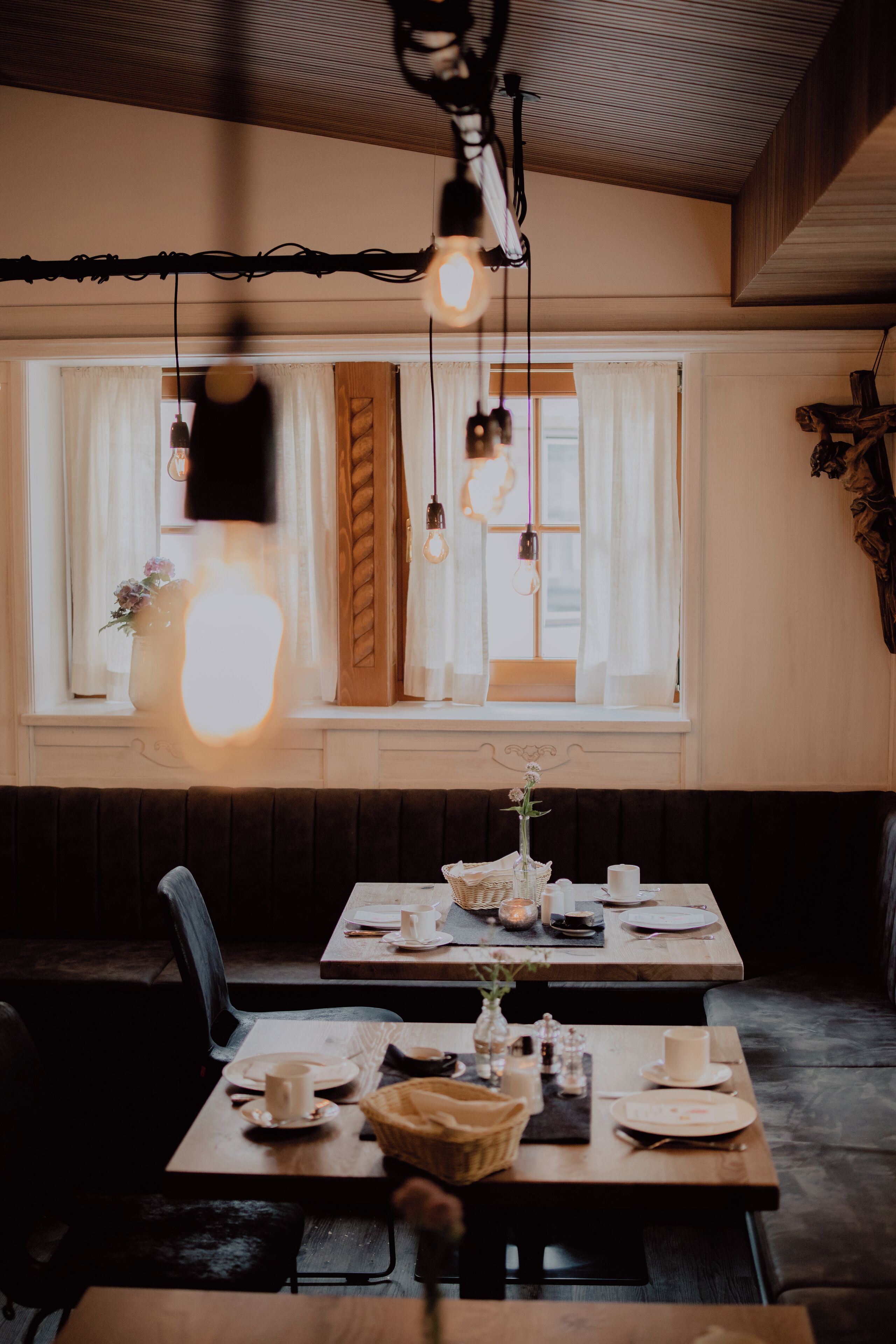 A cozy dining area with wooden tables and benches, a large window with curtains, and hanging light fixtures.