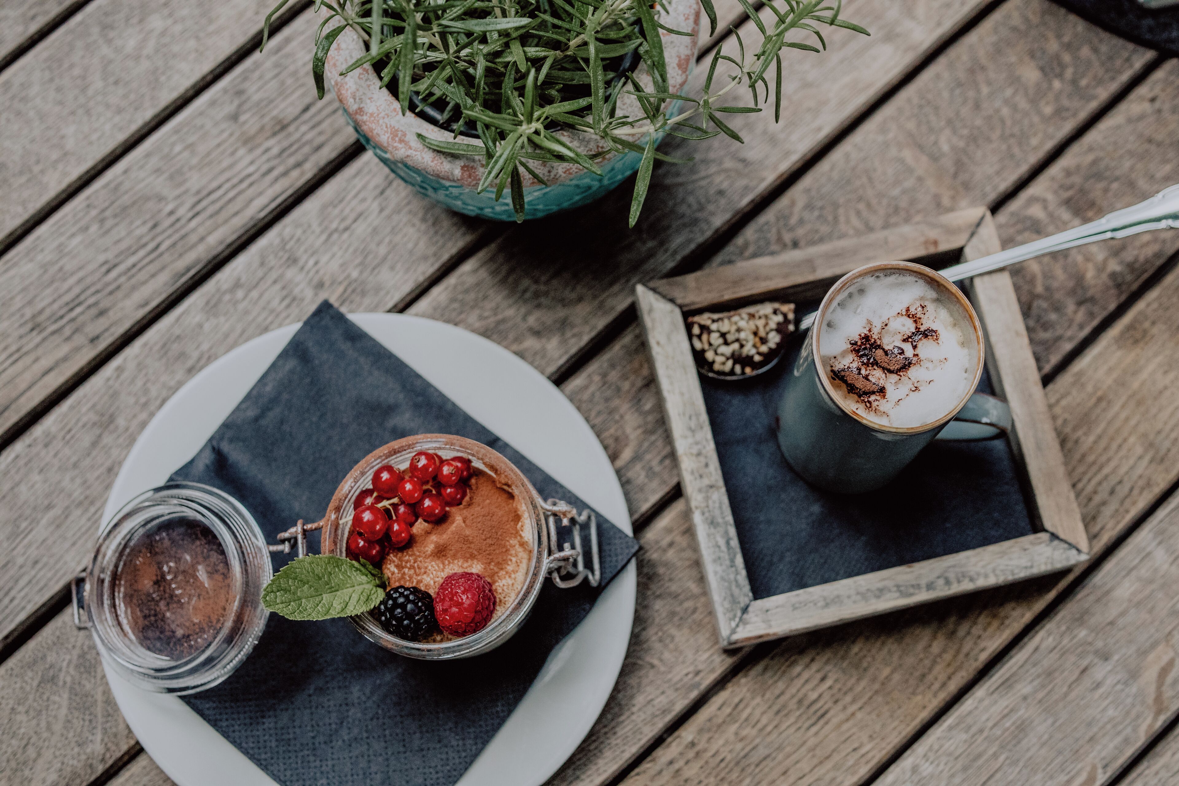 Une table en bois, dans un décor rustique, avec un bocal en verre contenant un dessert, une tasse de café et une plante en pot.