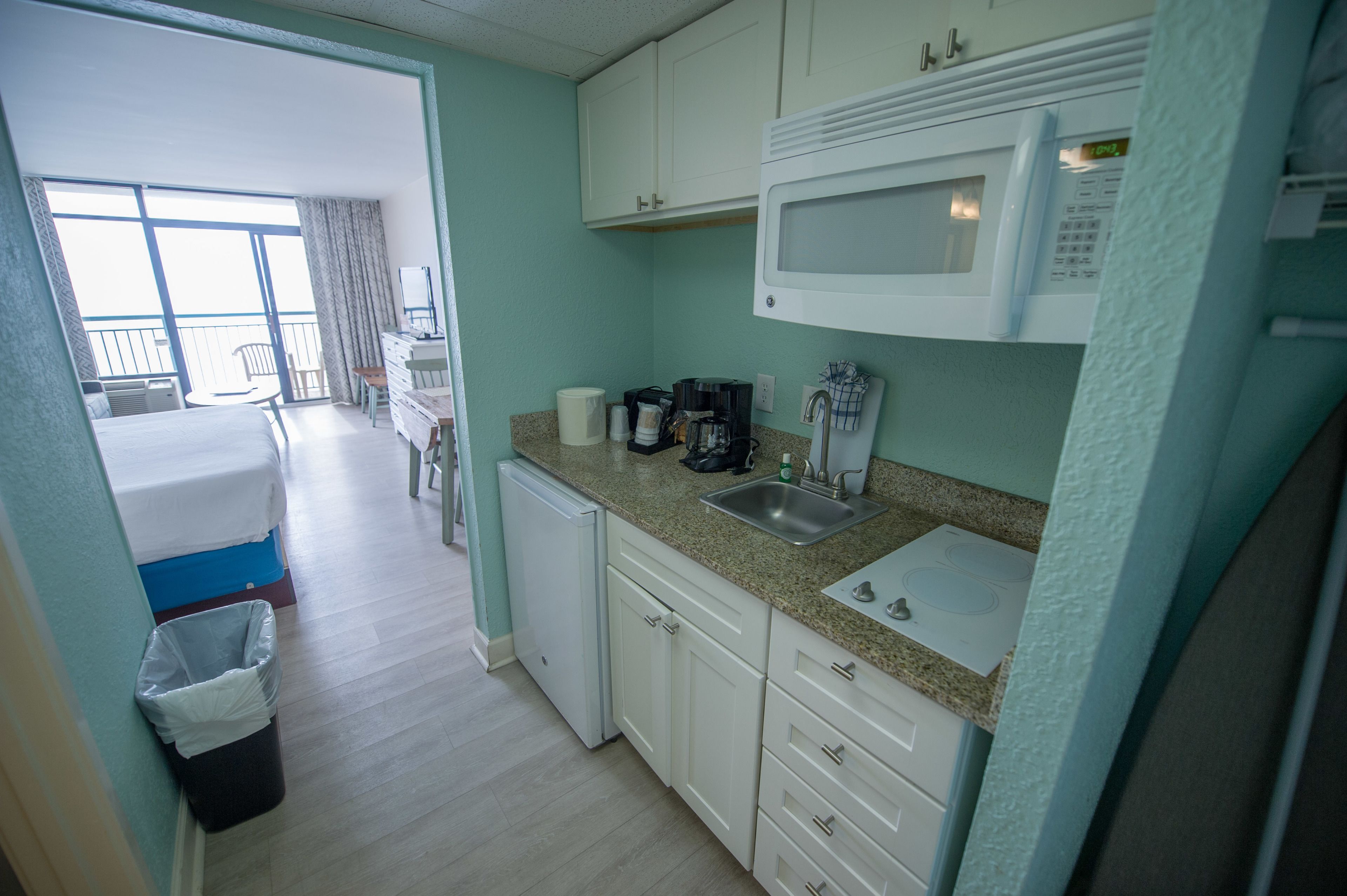 A compact hotel room with a kitchenette, a bed, a small bathroom, and a balcony with a view.