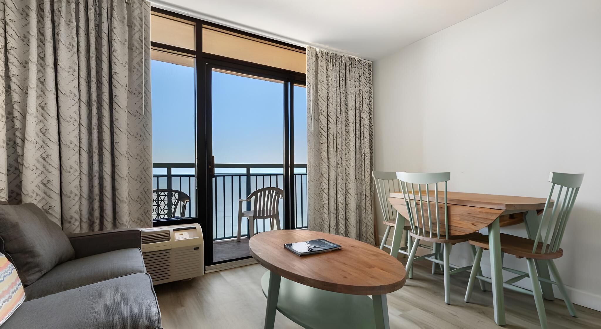 Ein modernes Wohnzimmer mit einer Couch, einem Holzesstisch mit Stühlen und einer großen Schiebeglastür, die auf einen Balkon führt.