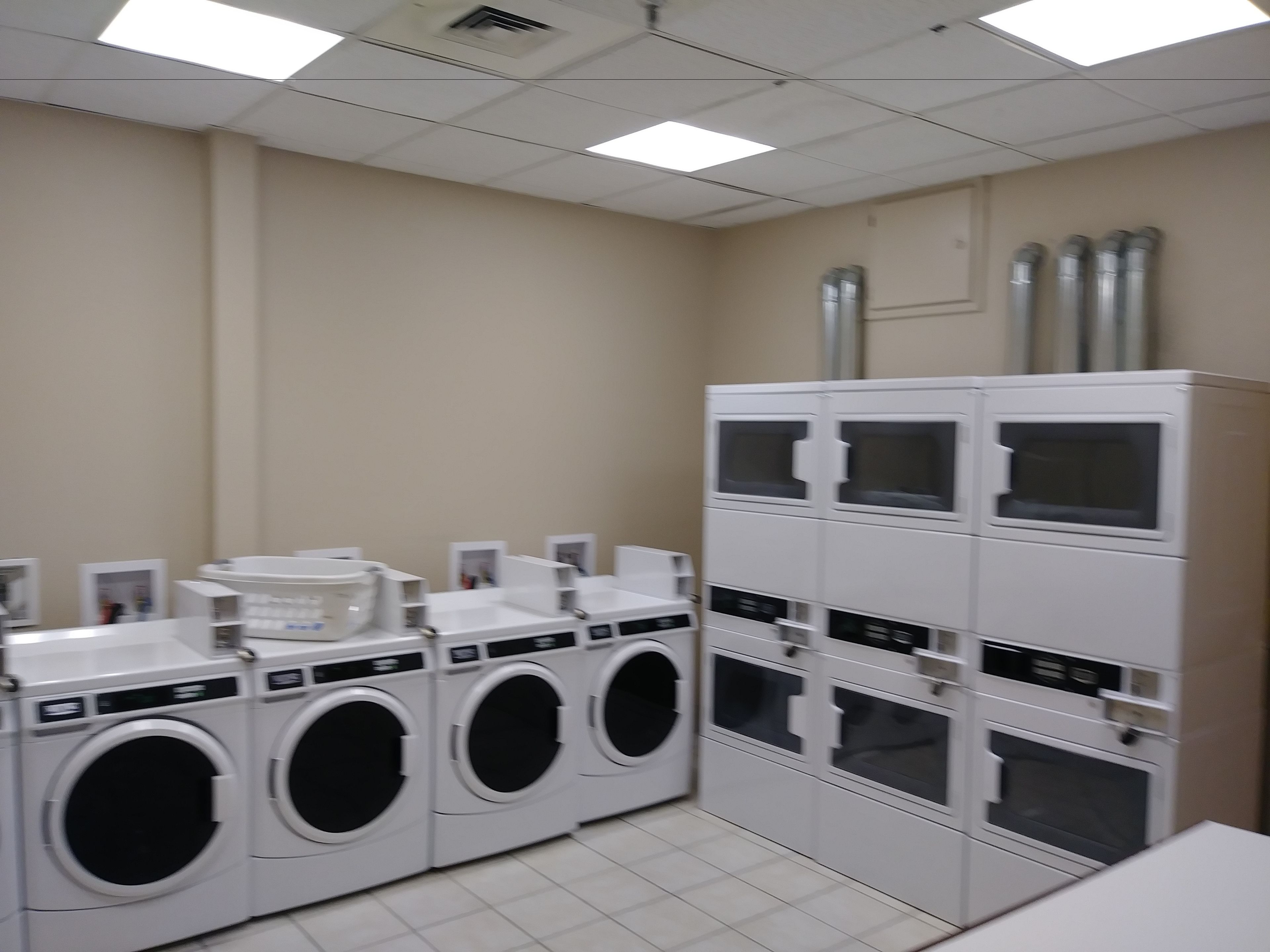A laundry facility with multiple washing machines and dryers, a tiled floor, and a ceiling with fluorescent lights.