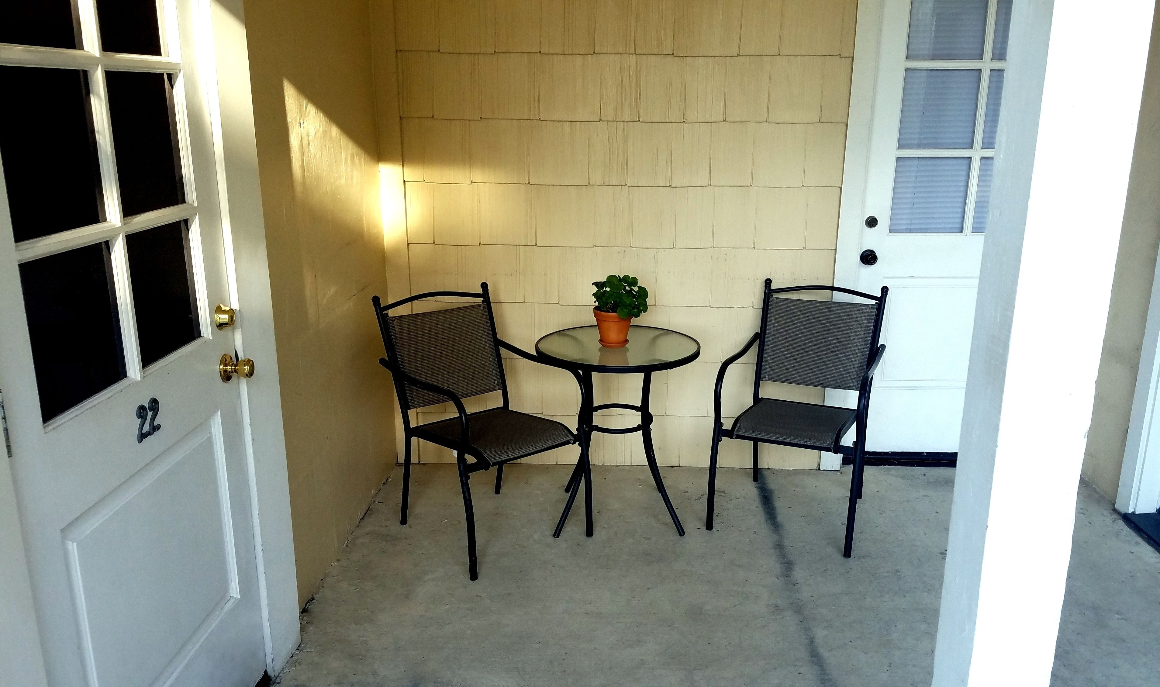 Un pequeño patio con dos sillas, una mesa, una planta en maceta y una puerta blanca con un ventanal.