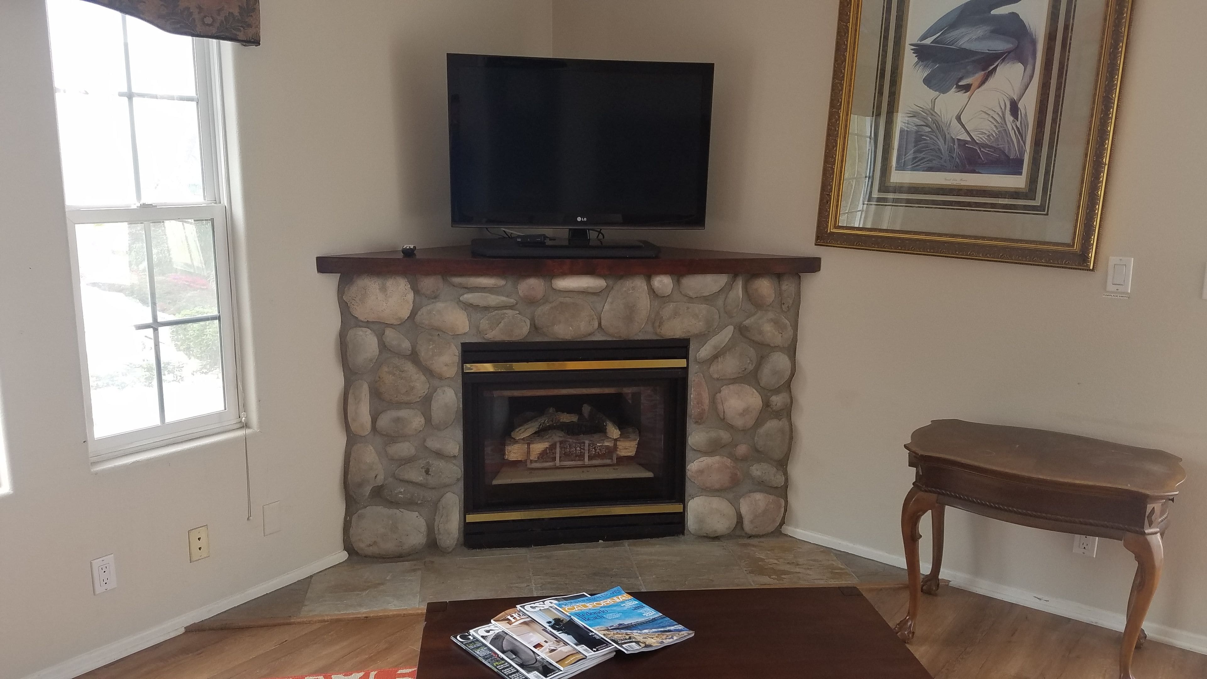 Een woonkamer met een stenen openhaard, een flatscreen televisie op een houten plank, een ingelijste foto aan de muur en een houten salontafel met tijdschriften.