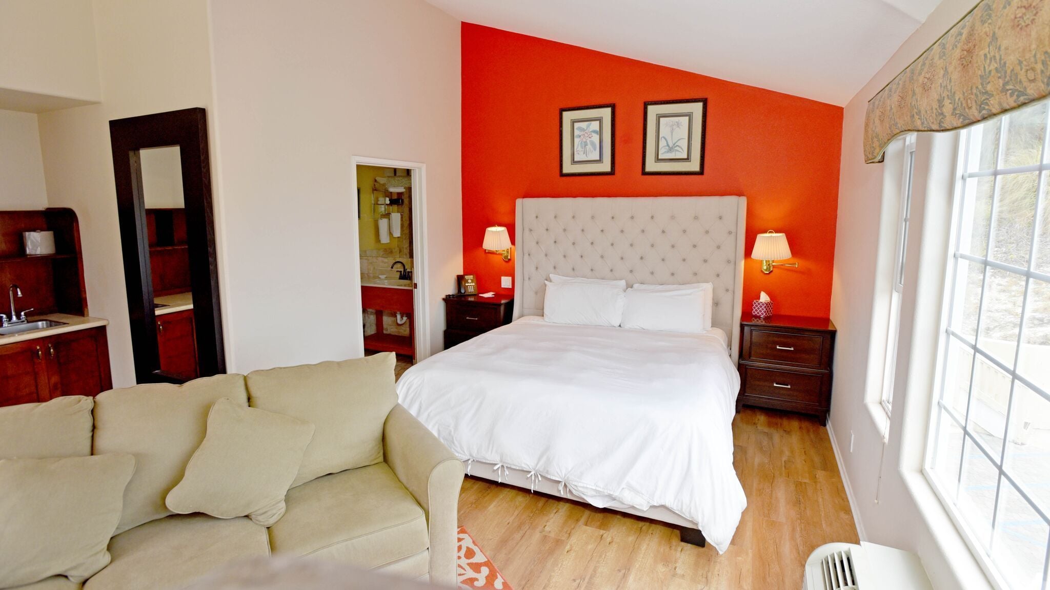 A hotel room with a large bed, a sofa, a nightstand, a mirror, and a window with a view.