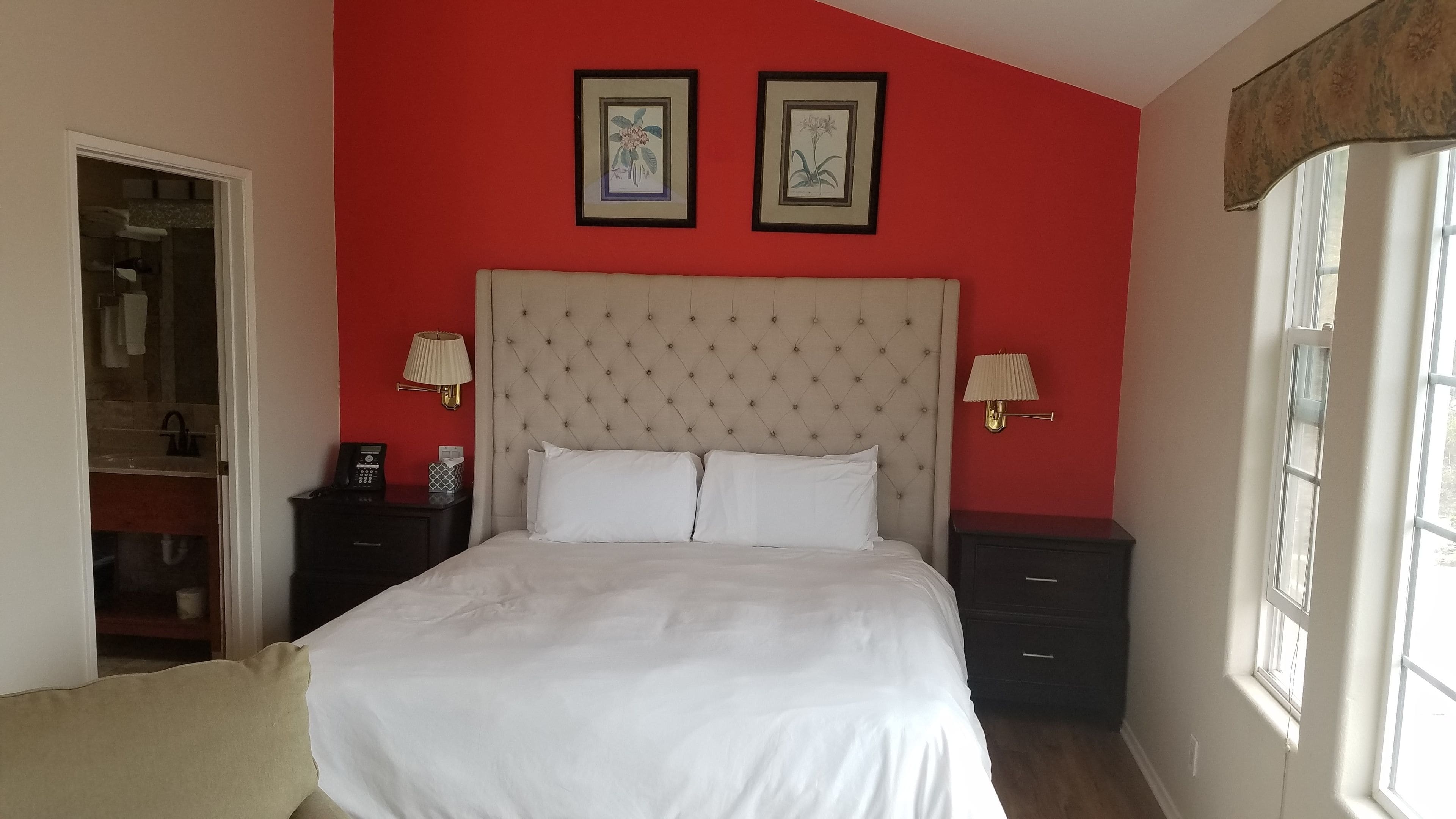 A bedroom with a tufted headboard, white bedding, two bedside lamps, and framed botanical prints on the wall.