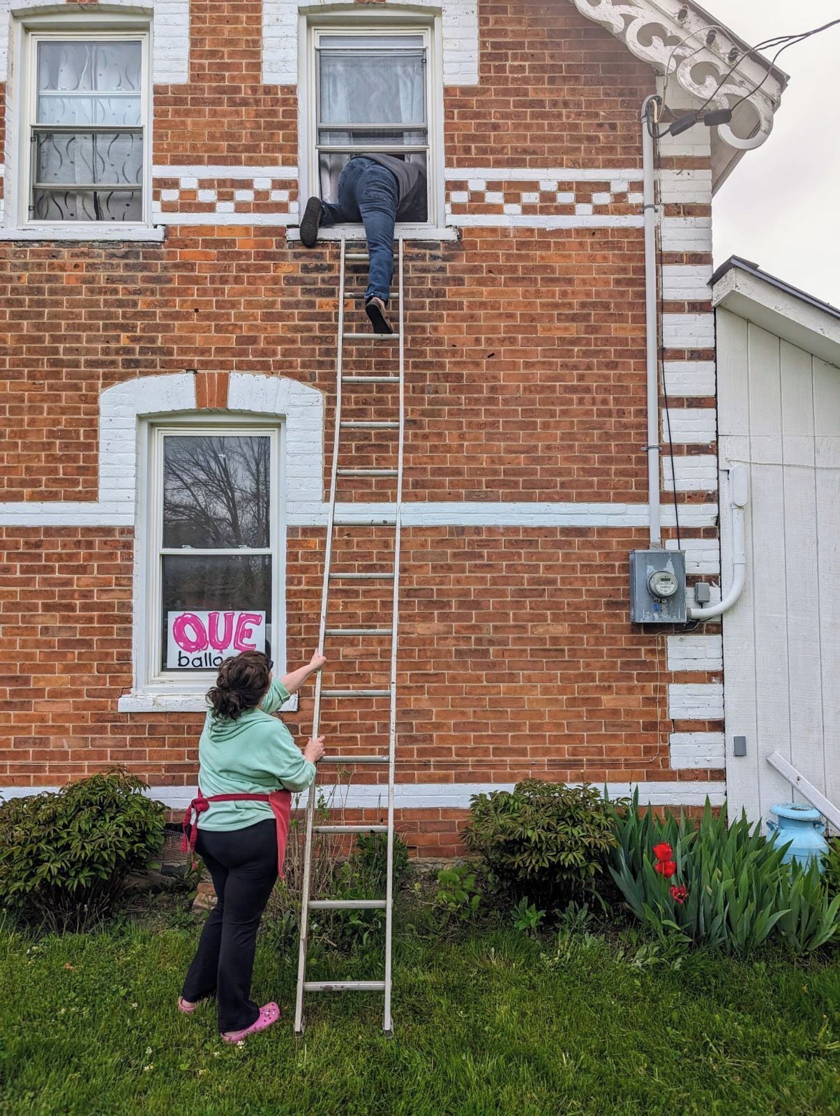 A person on a ladder against a brick building with a window labeled "QUE balloon."