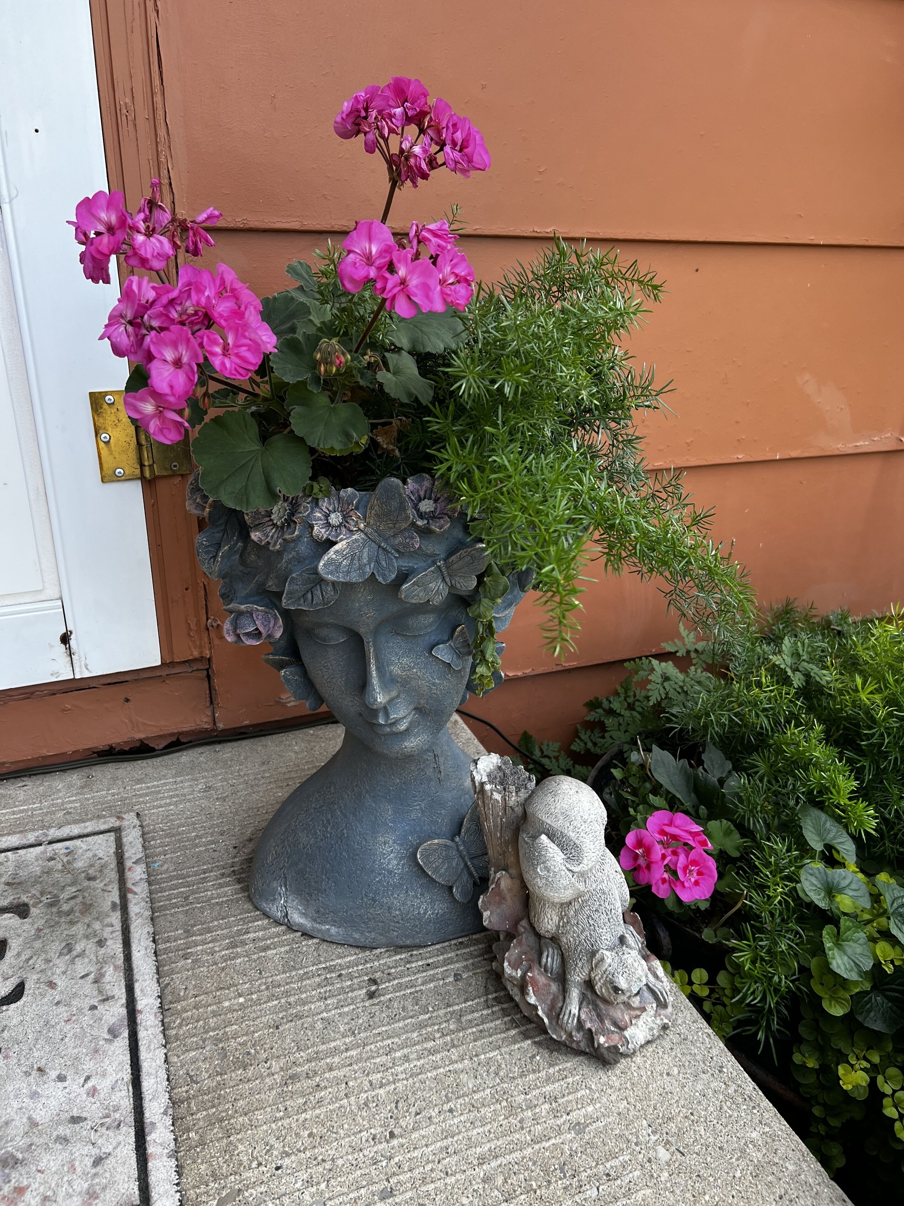 A concrete planter with a sculpted bust and a butterfly, containing pink flowers and green foliage, placed on a concrete surface against a building wall.