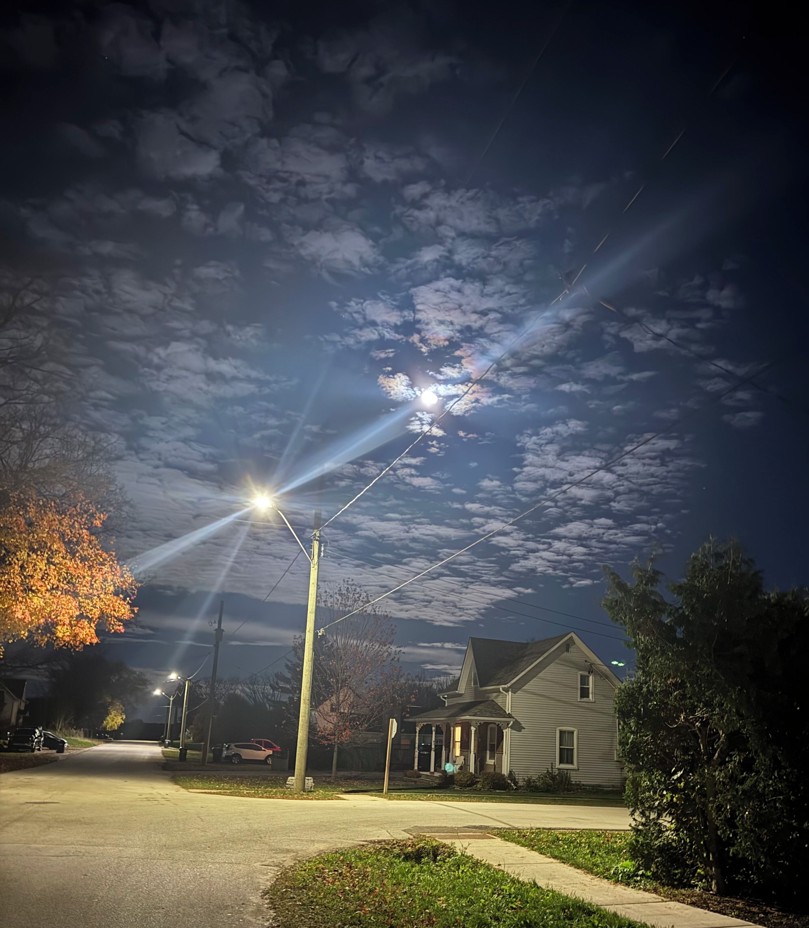Una calle residencial nocturna con farolas, casas y un cielo despejado con nubes.