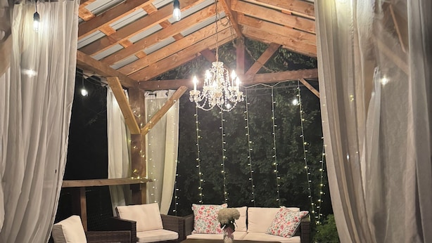 Un patio al aire libre con mobiliario de madera, cortinas blancas y luces colgantes.