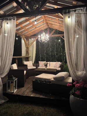 An outdoor patio with wooden furniture, white curtains, and string lights. - Emily's Place B&B (Clearview)