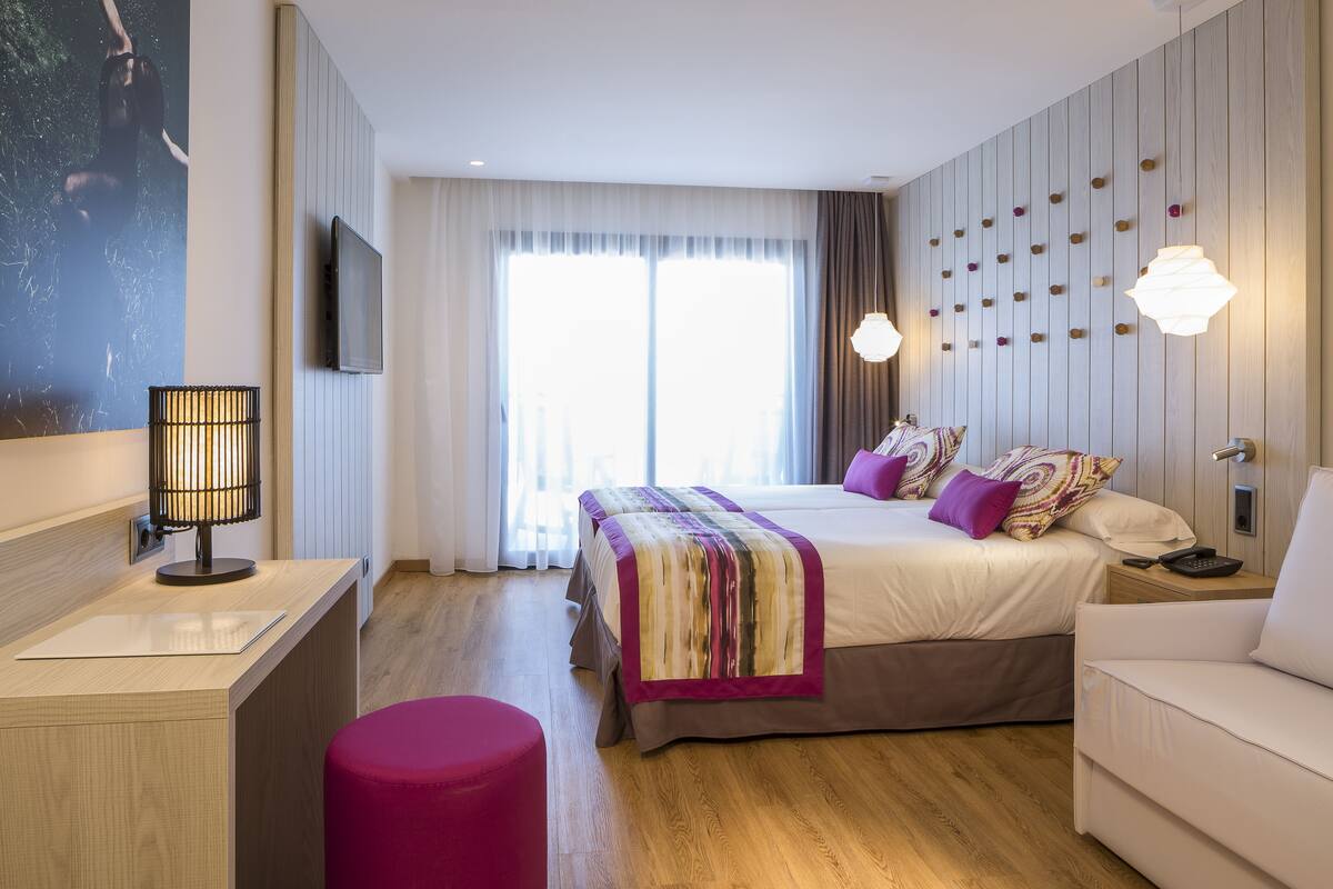 Una habitación de hotel moderna con una cama grande, un escritorio, un taburete rosa y un televisor montado en la pared.