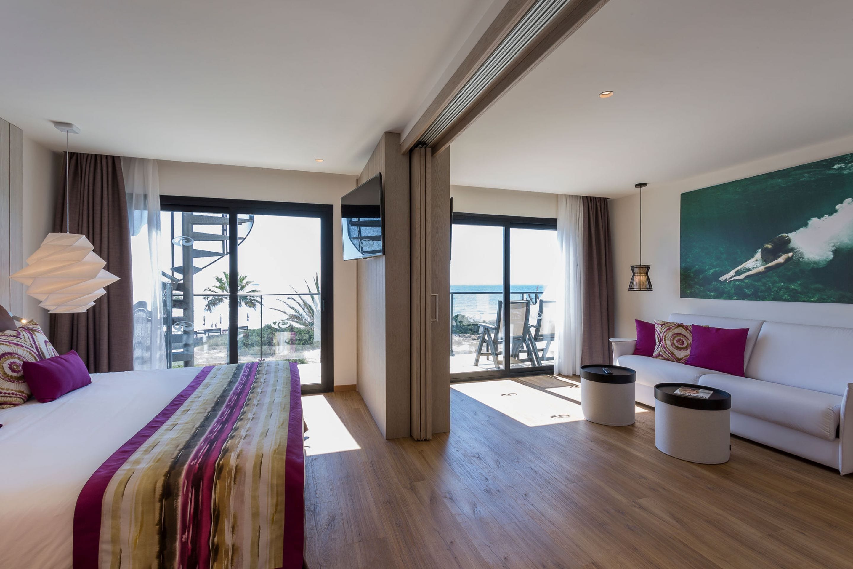 A modern hotel room with a large bed, a sofa, a balcony with a view, and a painting on the wall.