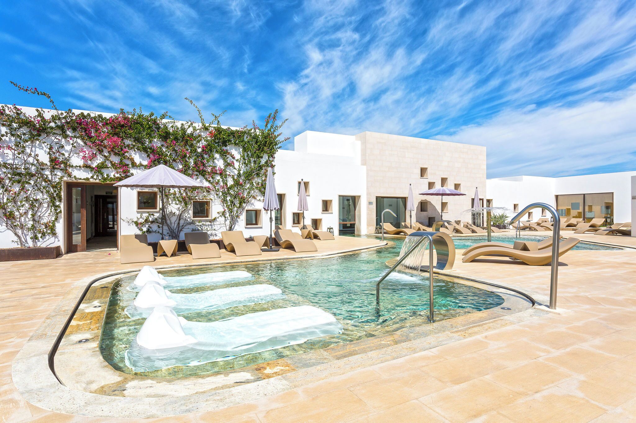 A pool area with lounge chairs and umbrellas, a clear sky, and a building with white walls and pink flowers.