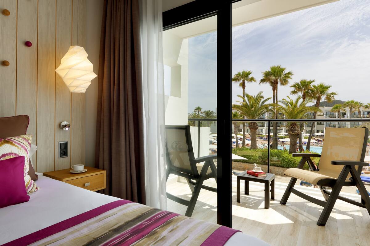Habitación de hotel con cama, mesita de noche, lámpara y vista a la piscina y palmeras.