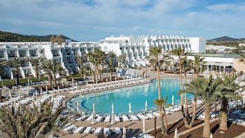 Un resort con una gran piscina rodeada de tumbonas y palmeras, edificios blancos modernos y un cielo azul despejado.