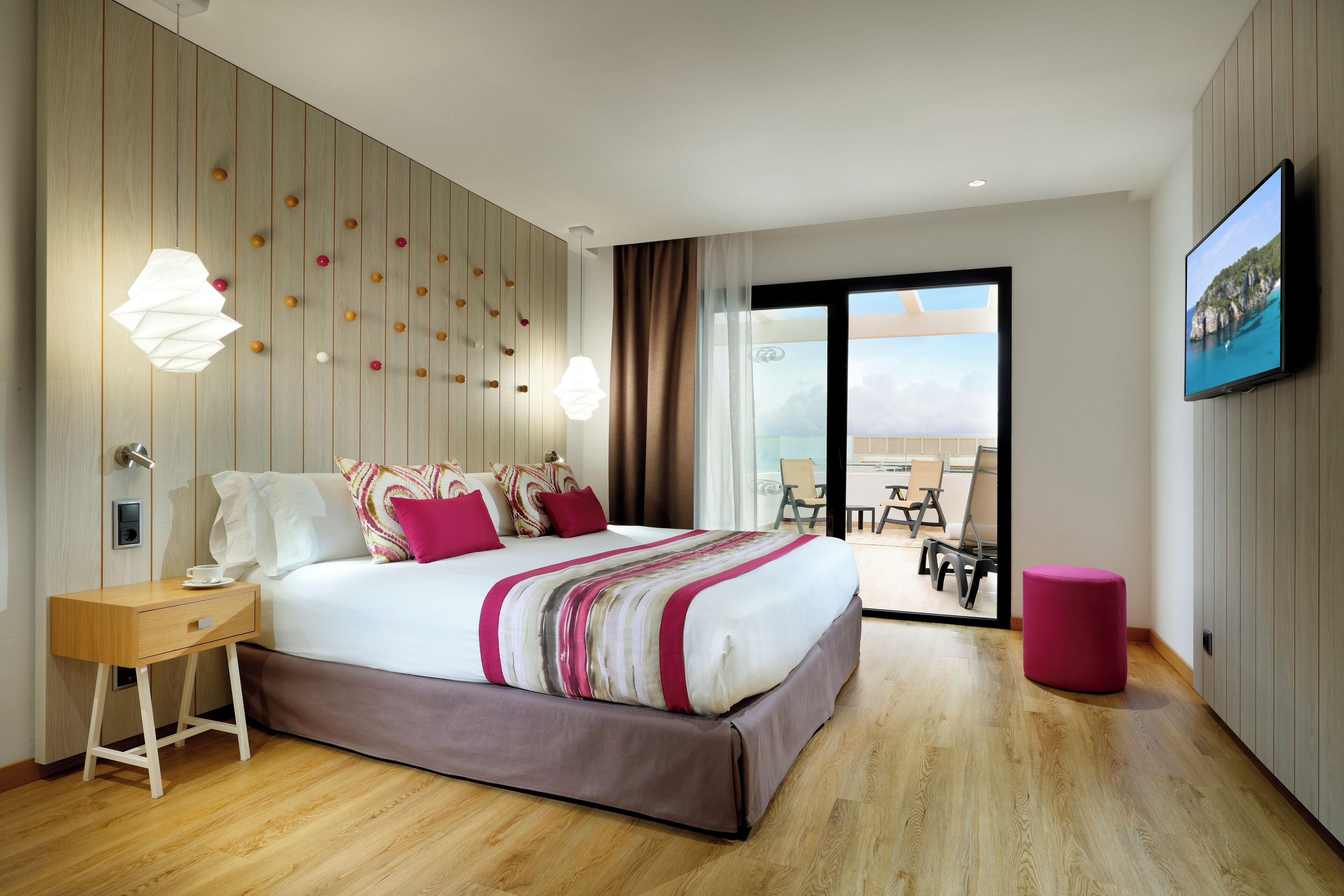 A modern hotel room with a large bed, a wooden nightstand, a pink ottoman, and a balcony with a view of the sea.