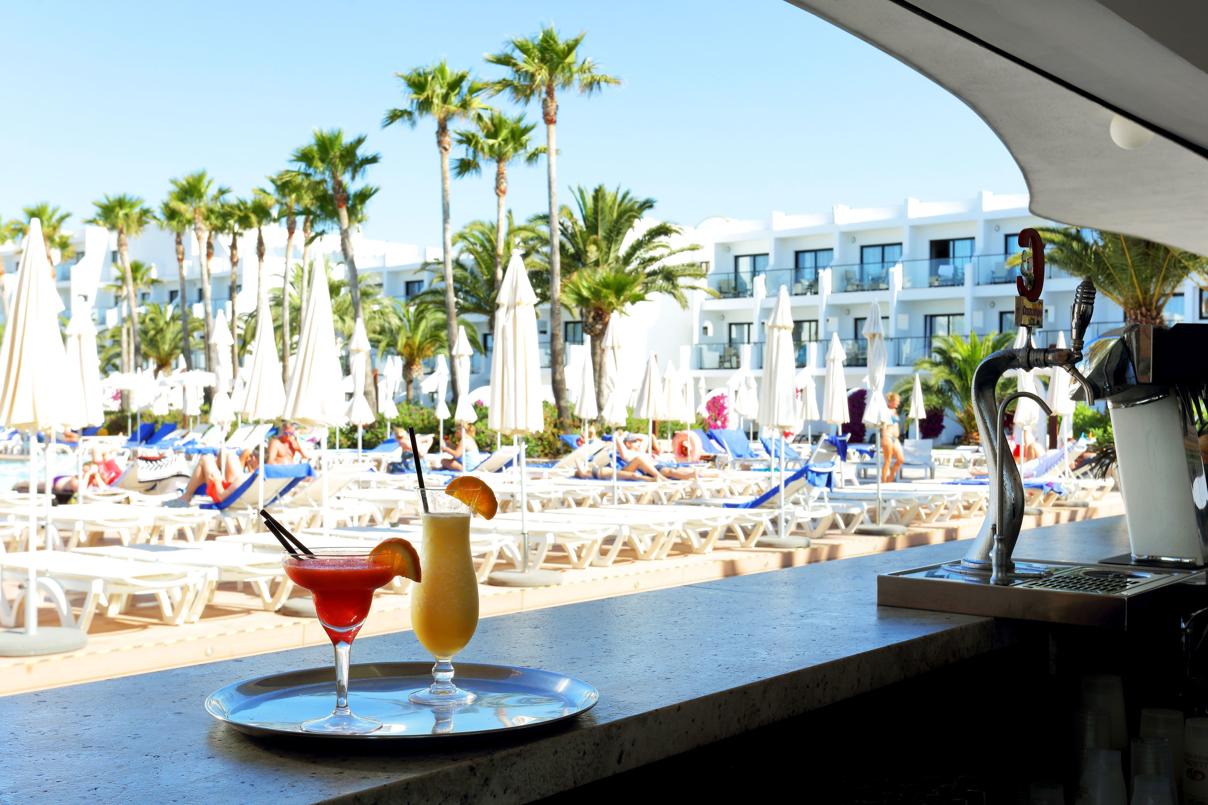 Eine Strandbar mit einem Cocktail auf einem Tablett, mit Blick auf einen Poolbereich mit Liegestühlen und Sonnenschirmen.