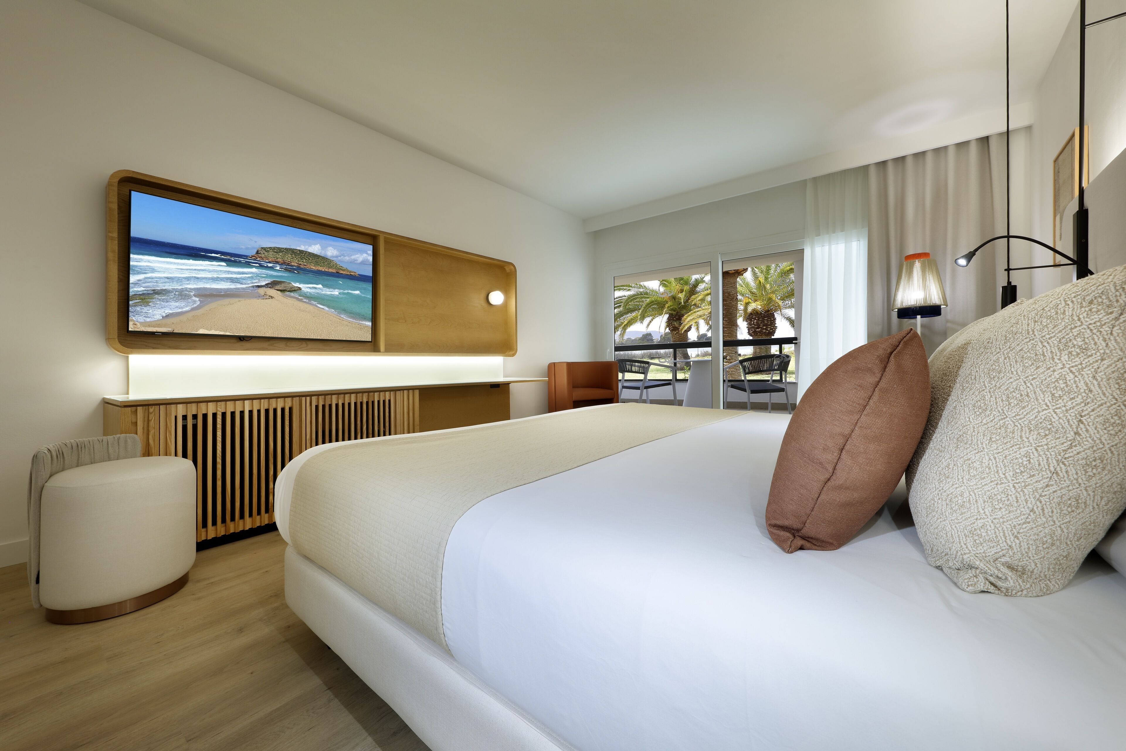 Une chambre d’hôtel moderne avec un grand lit, un téléviseur à écran plat fixé au mur, une table de chevet en bois, un petit pouf rond et une vue sur la plage par la fenêtre.