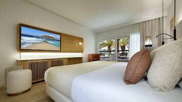 Une chambre d’hôtel moderne avec un grand lit, un téléviseur à écran plat fixé au mur, une table de chevet en bois, un petit pouf rond et une vue sur la plage par la fenêtre.