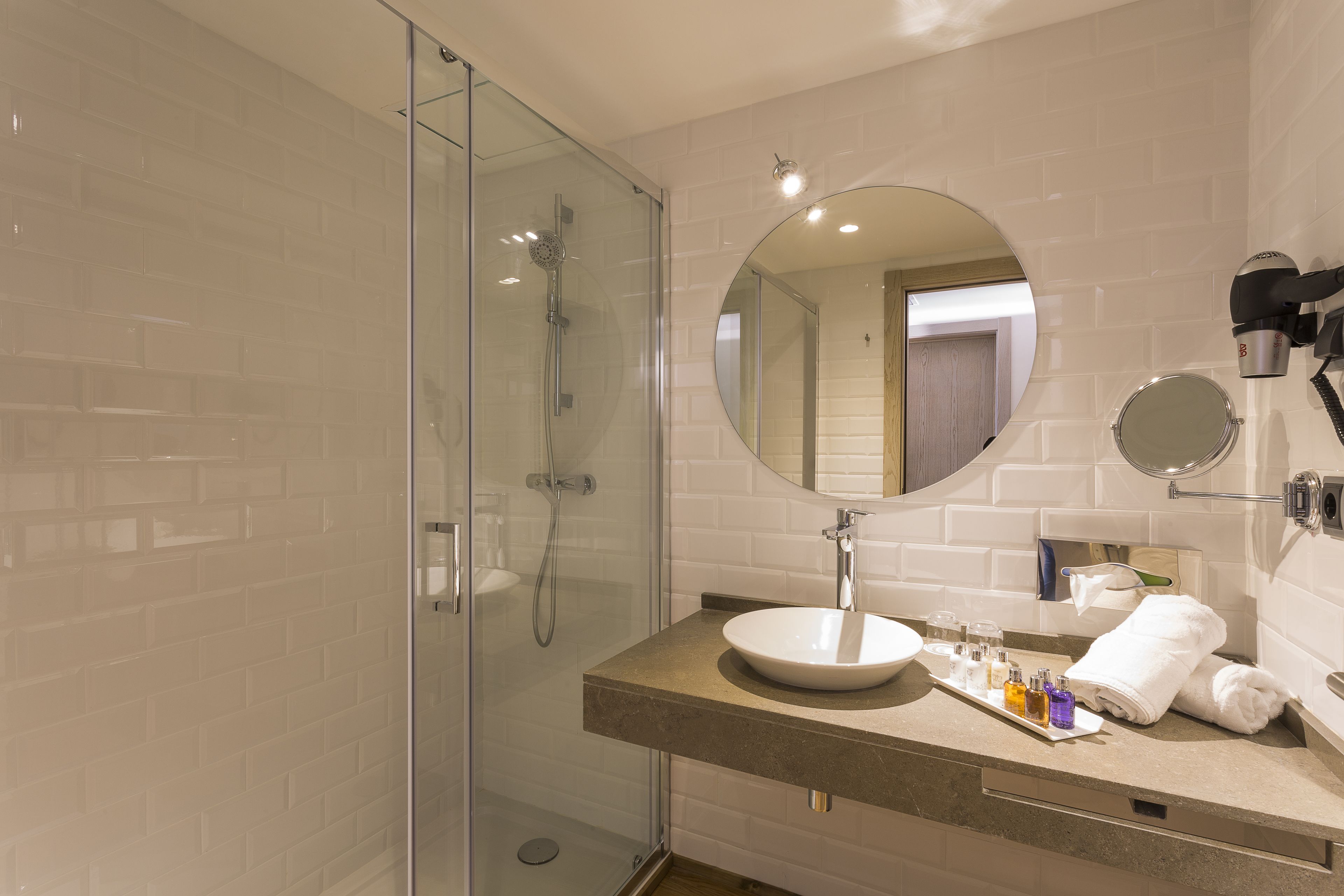 Un baño moderno con ducha envuelta en vidrio, un espejo redondo y una encimera color beige con artículos de aseo.