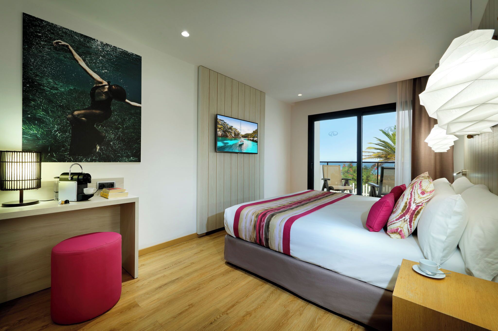 A modern hotel room with a large bed, a desk with a computer, a red ottoman, a TV on the wall, and a view of the sea through the window.