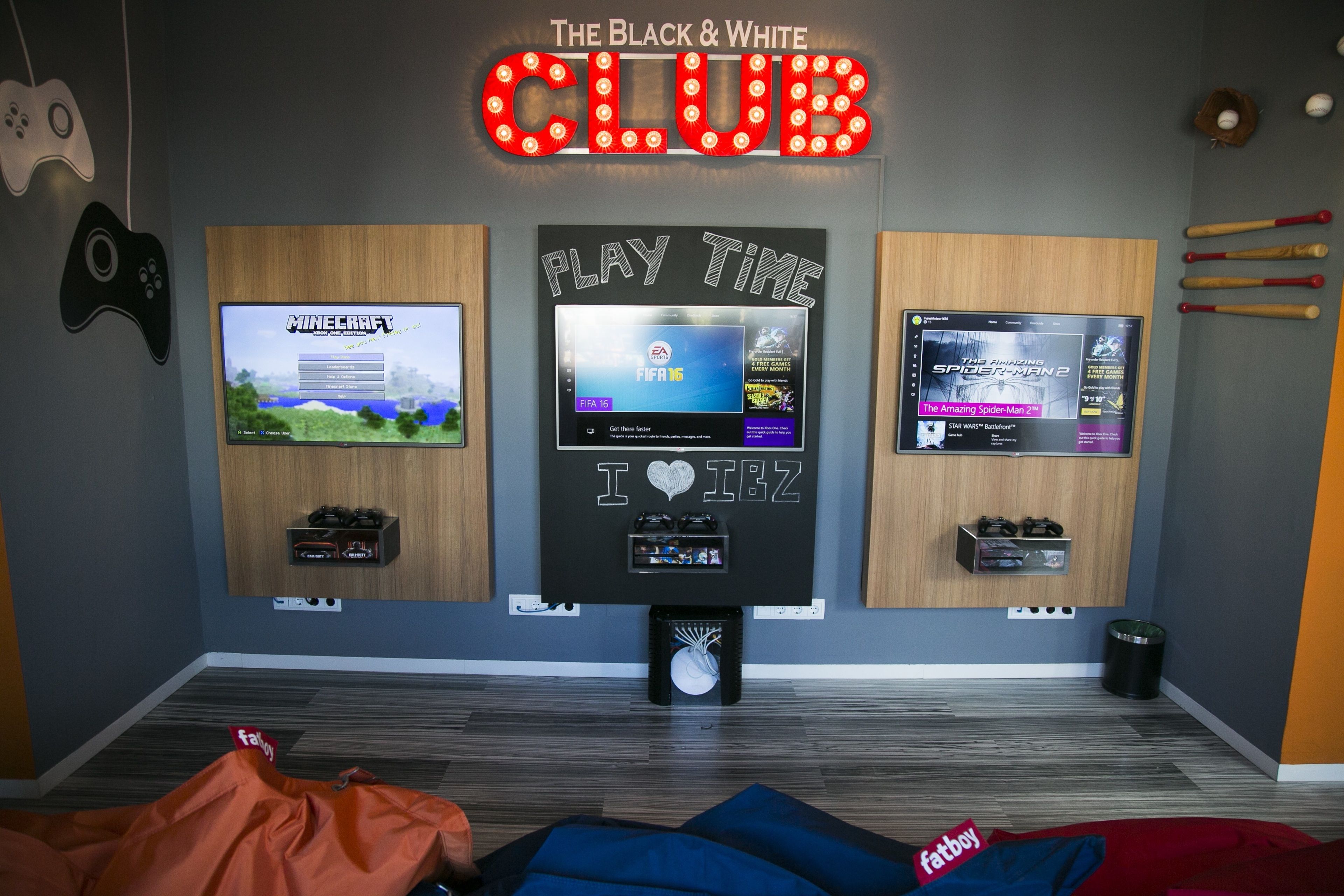 A gaming room with three TVs displaying different games, a neon sign reading "The Black & White CLUB", and various gaming accessories.