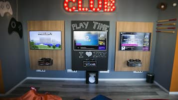 A gaming room with three TVs displaying different games, a neon sign reading "The Black & White CLUB", and various gaming accessories.