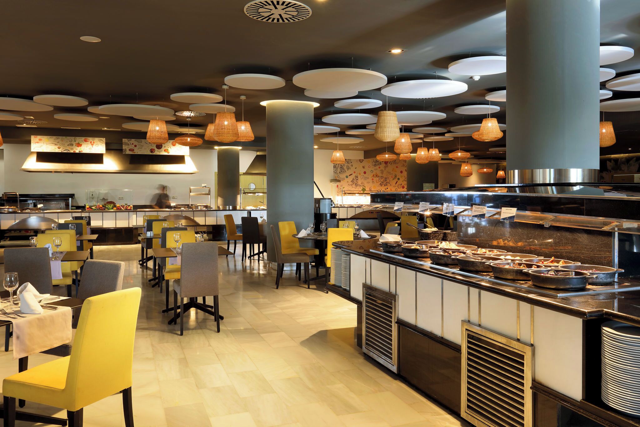 Ein modernes Restaurant mit einem zentralen Buffet, gelben und grauen Stühlen und Pendelleuchten.