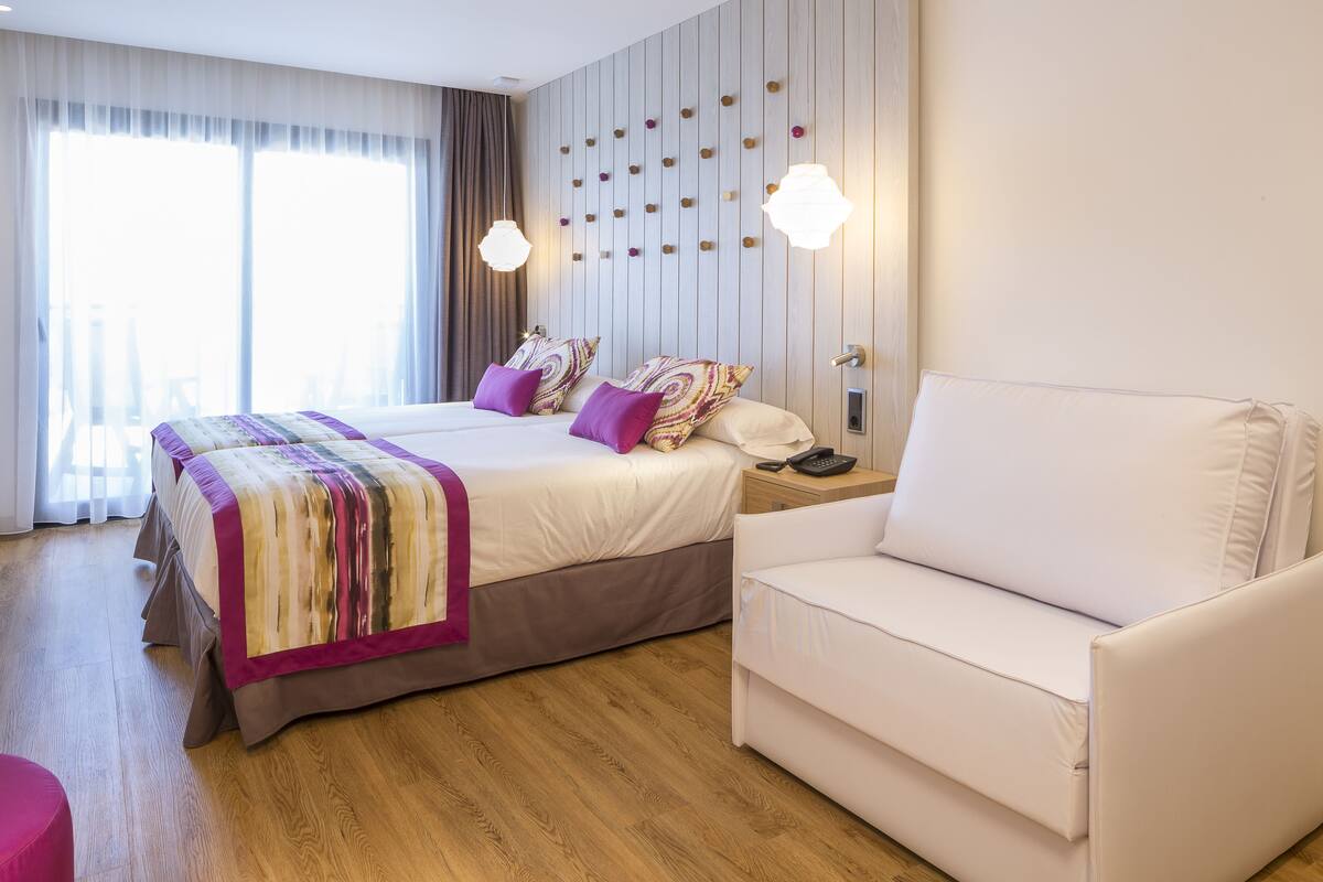 Habitación de hotel con una cama grande, un sillón blanco y piso de madera.