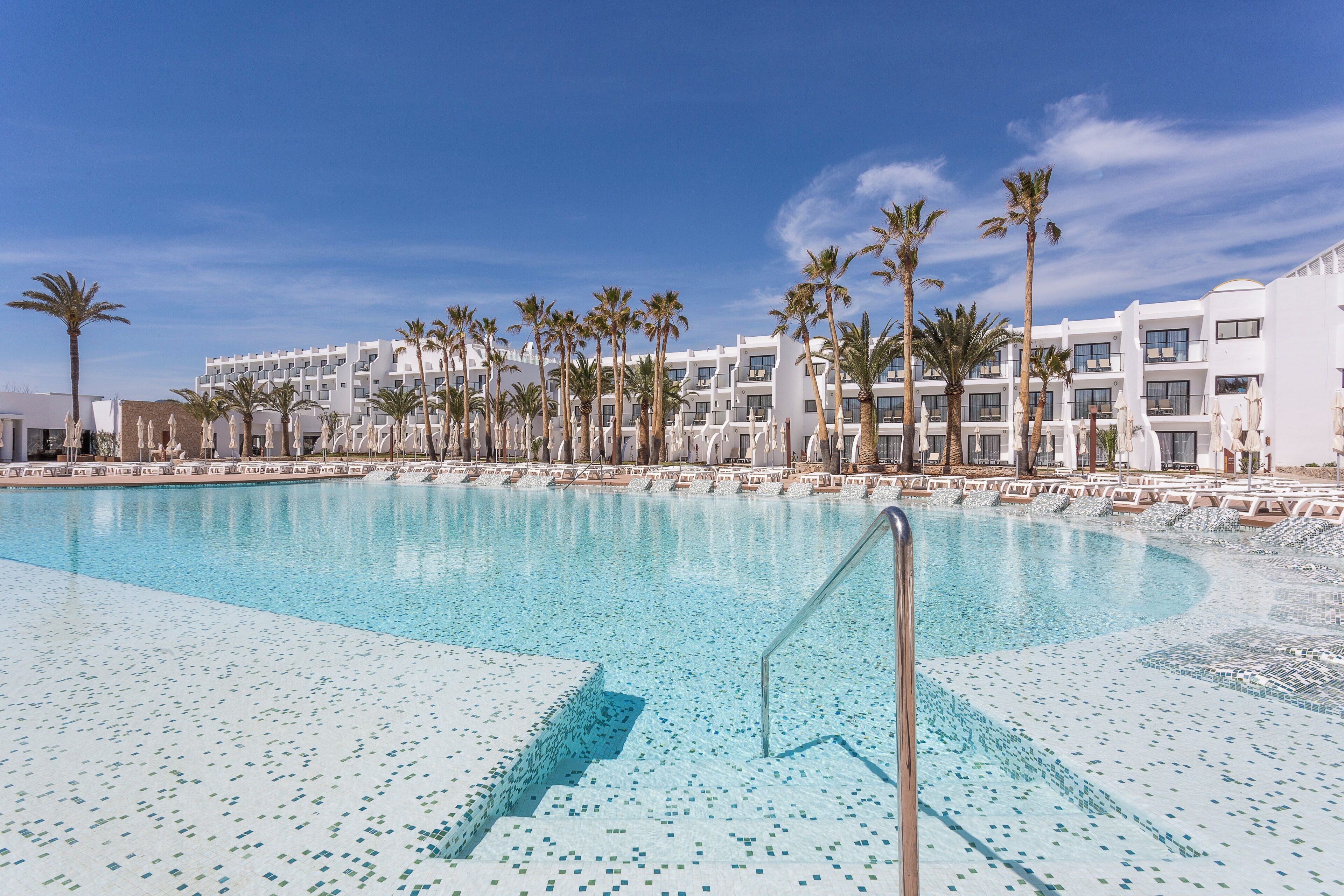 Une piscine dotée d’une terrasse carrelée, entourée de palmiers et bordée d’un immeuble d’hôtel à plusieurs étages.