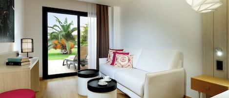A modern hotel room with a white sofa, a red ottoman, a wooden desk with a lamp, and a view of the outdoors.