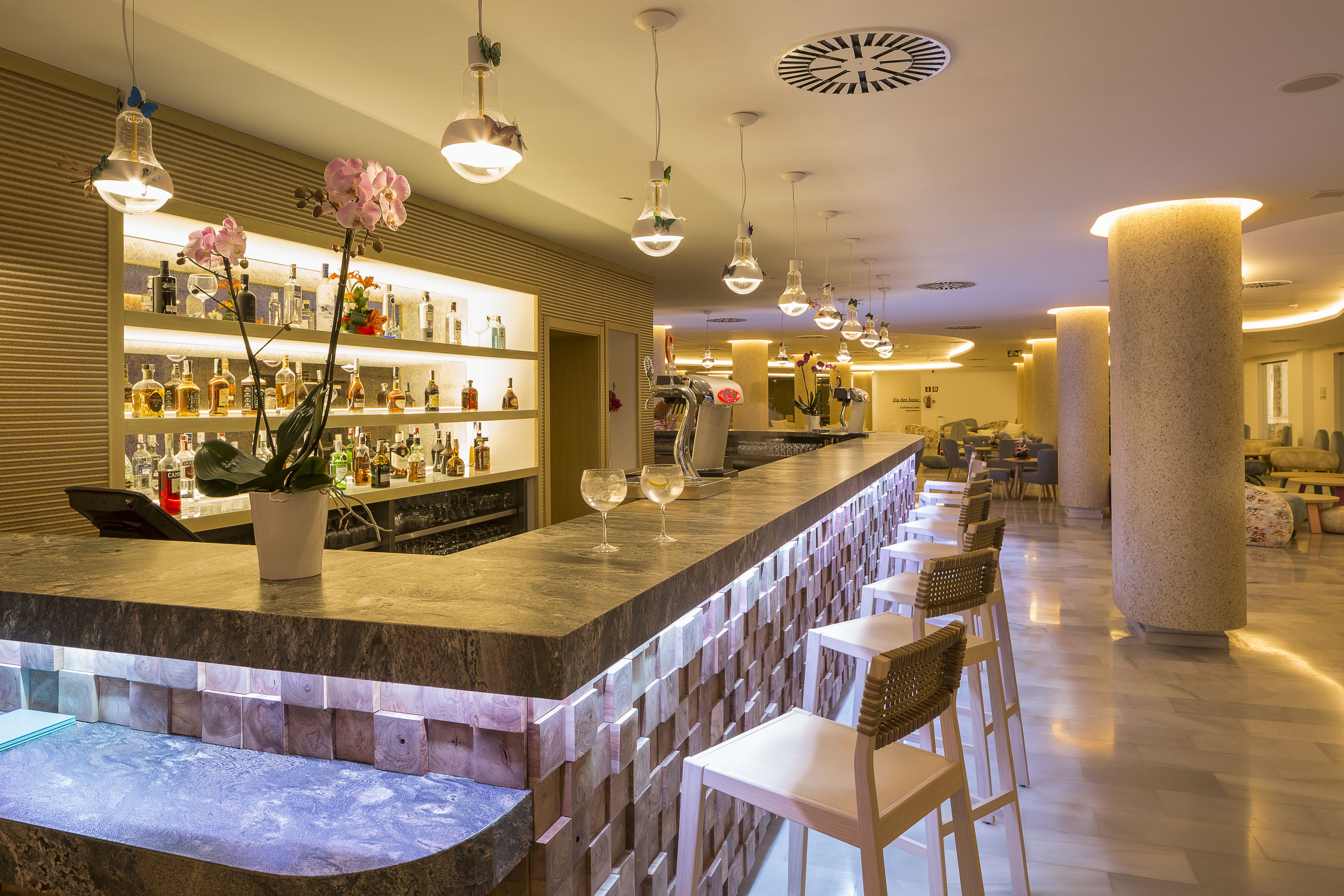 Una zona de bar con mostrador de mármol, varias lámparas colgantes y una variedad de botellas y copas. Hay varios taburetes y una zona de estar con mesas y sillas al fondo.