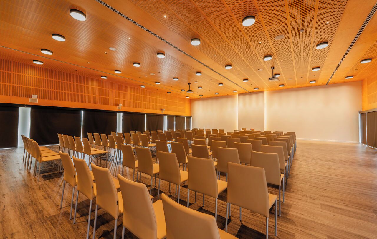 Een vergaderruimte met rijen stoelen, een groot scherm en een houten plafond met ronde lampen.