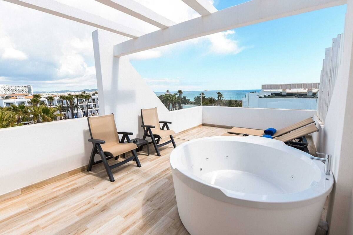 Un balcón con una bañera grande, dos sillas y vistas al mar y edificios.