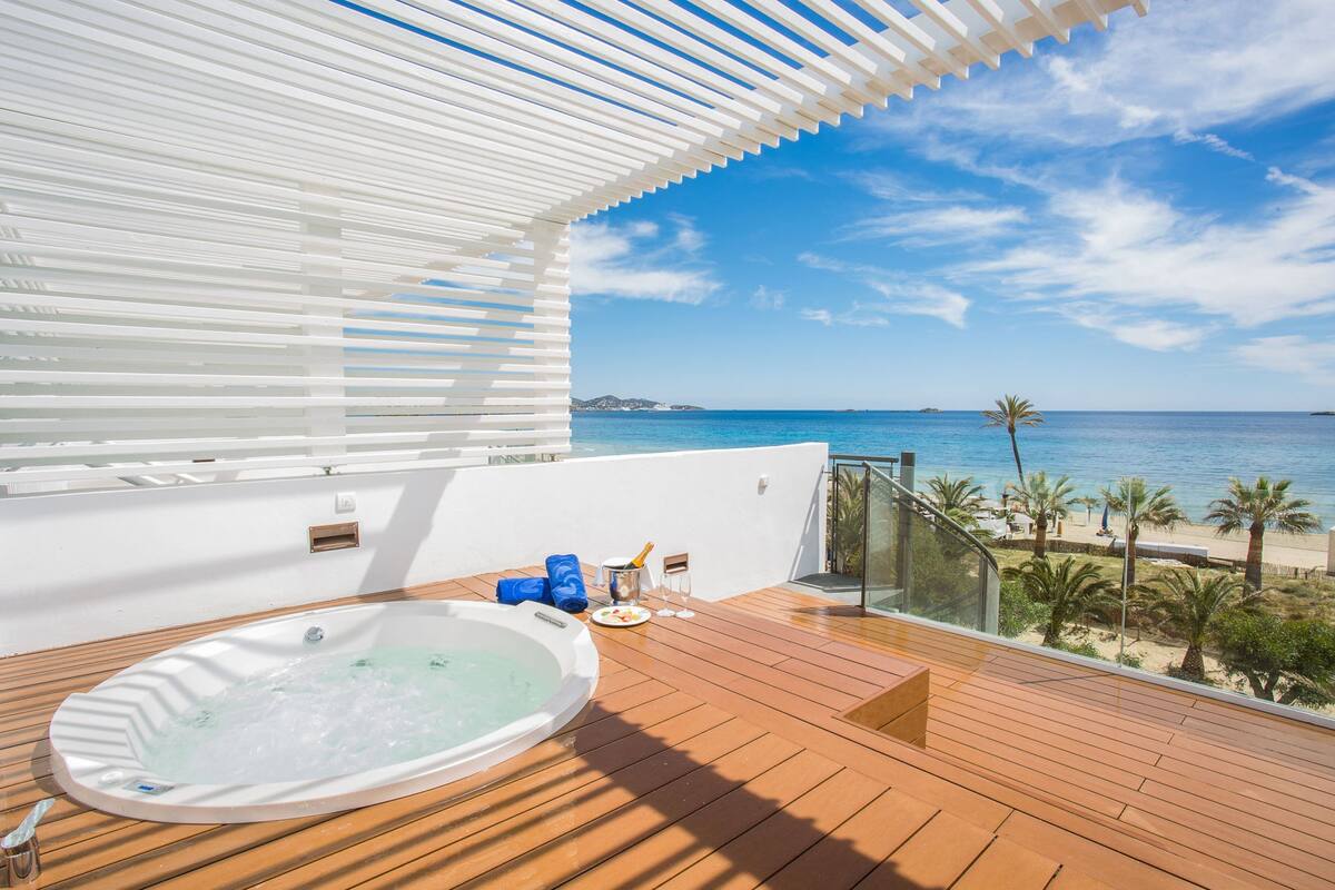 Una terraza de madera con un jacuzzi, un balcón con paredes de vidrio y vistas al mar.