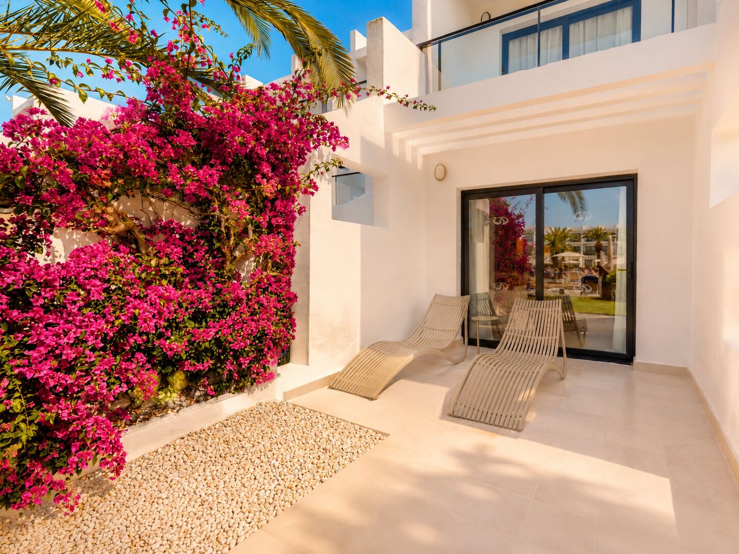 Ein moderner Außenbereich mit Sitzgelegenheit, eine Glastür führt zu einem Pool, umgeben von weißen Wänden und rosa Bougainvillea.