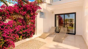 A modern outdoor seating area with a glass door leading to a pool, surrounded by white walls and pink bougainvillea.
