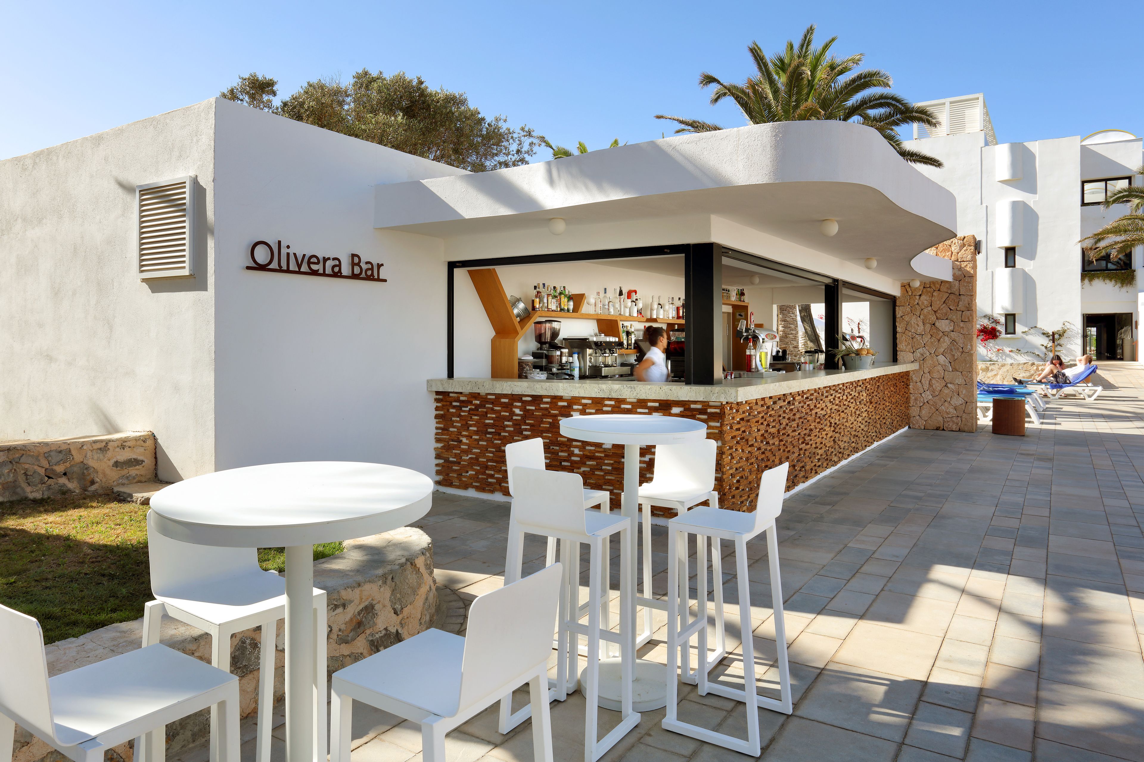 An outdoor bar area with white tables and chairs, a stone wall, and a clear sky.