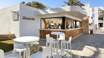 An outdoor bar area with white tables and chairs, a stone wall, and a clear sky.