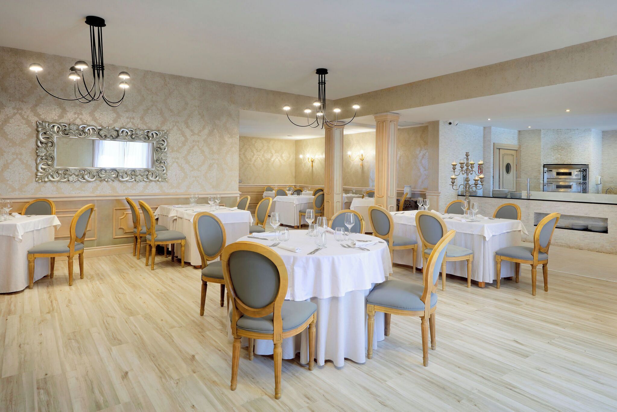 A dining area with round tables, chairs, chandeliers, and a large mirror on the wall.