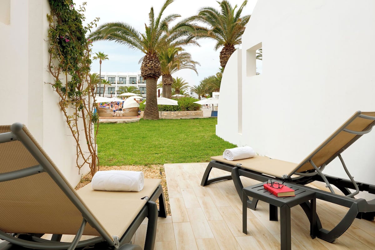 Un área de descanso al aire libre iluminada por el sol, con dos sillones, una mesita con un libro y vasos, y vistas a una piscina y palmeras.