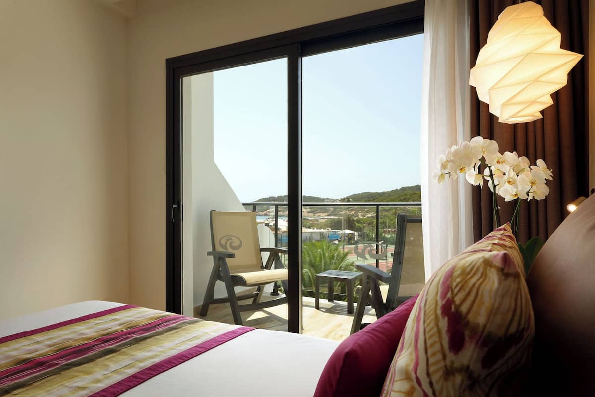 Habitación de hotel con cama, una silla, una mesita y un balcón con vistas al paisaje.
