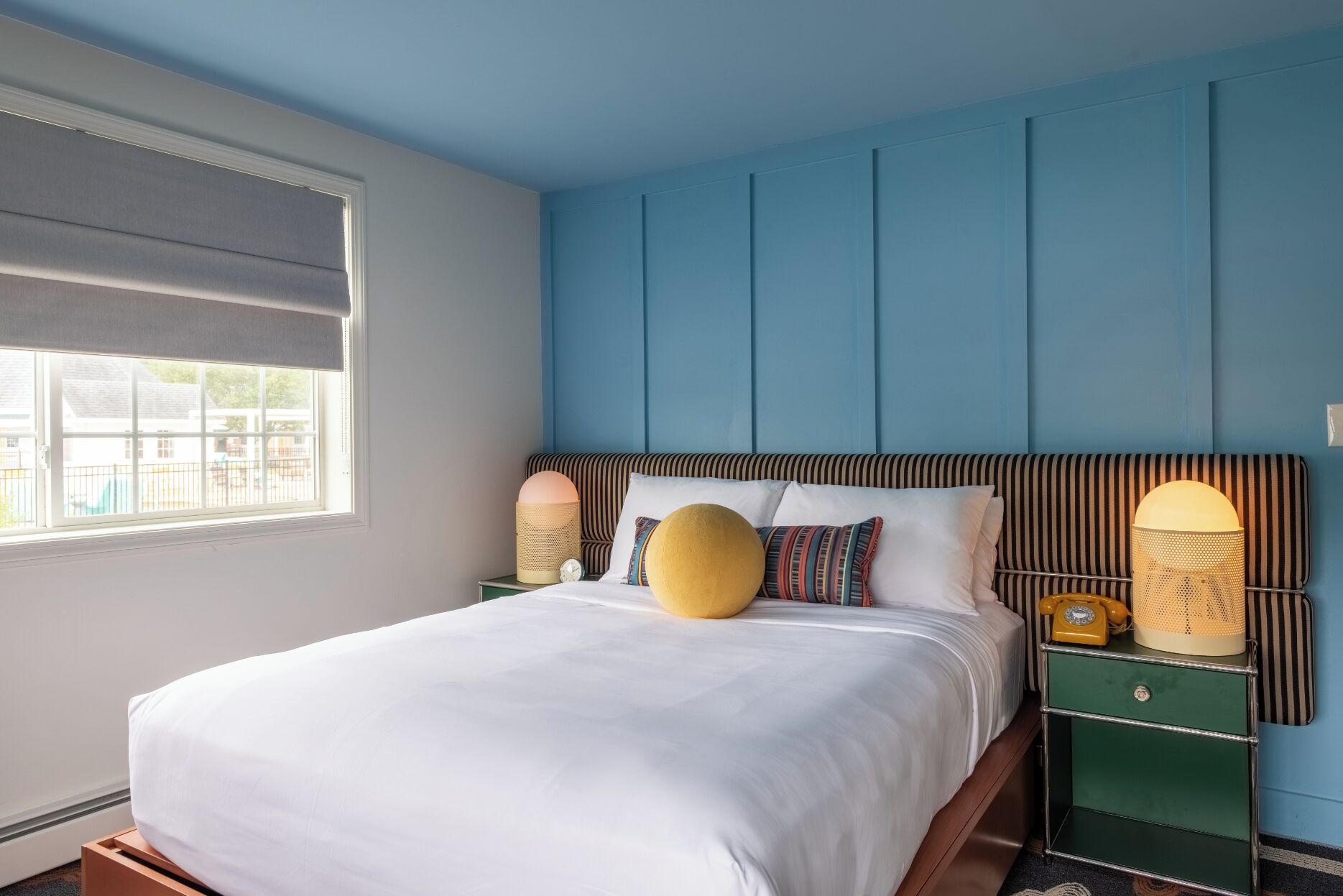 A bedroom with a bed, a window, a bedside table with a lamp, and a wall-mounted telephone.