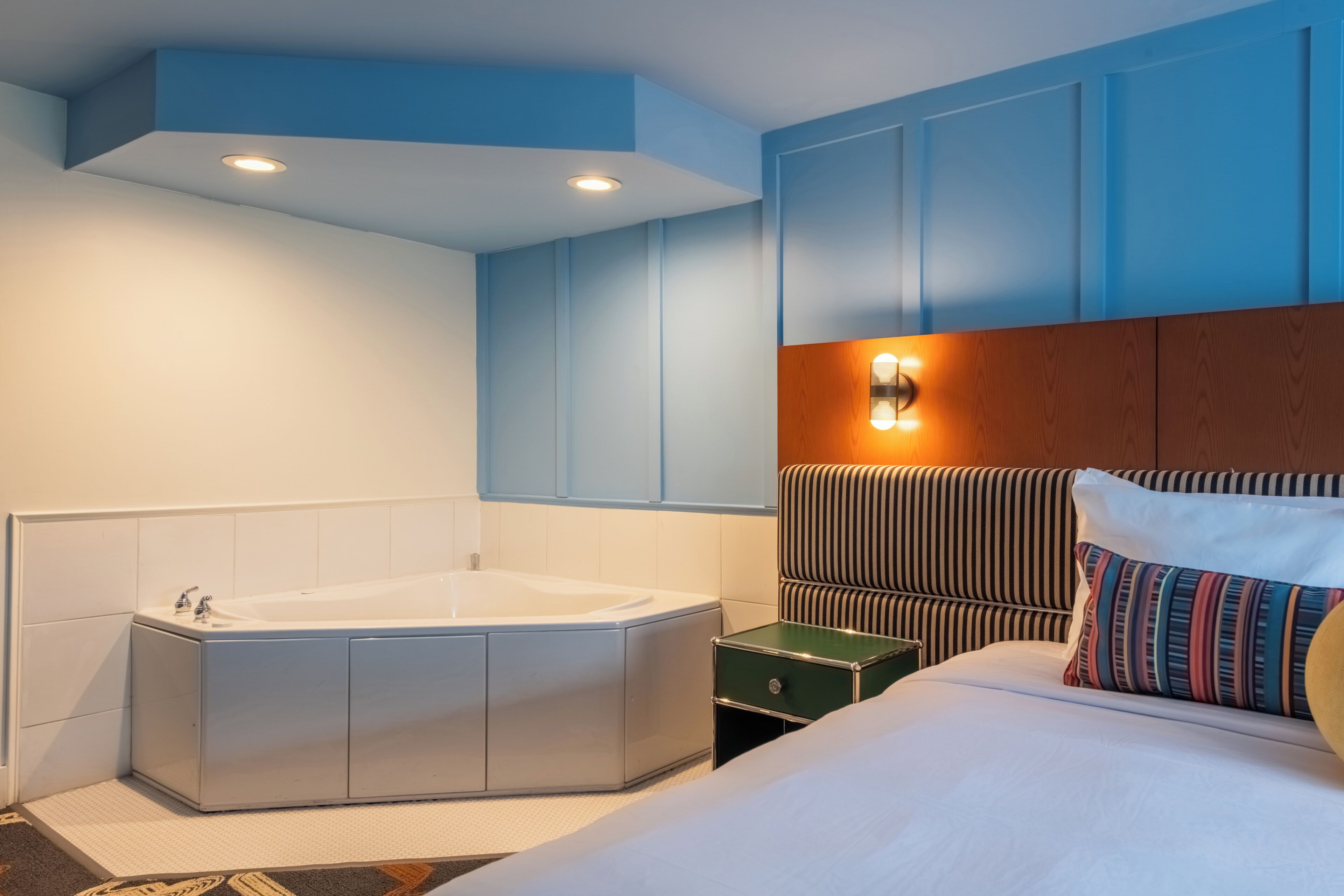 A hotel room with a large bathtub, a bed with striped pillows, and a nightstand with a lamp.