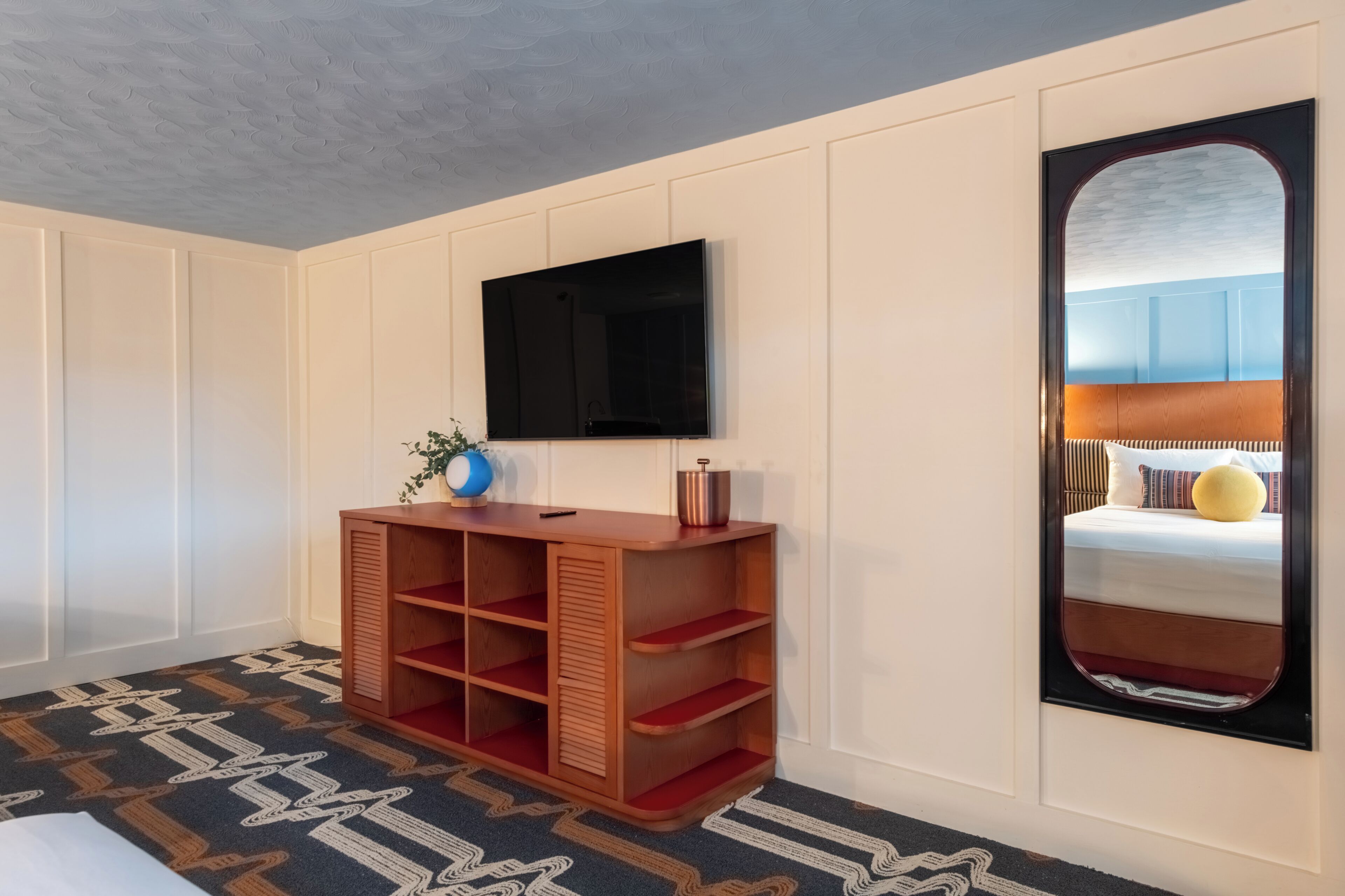 Quarto de hotel com televisão na parede, mesa consola de madeira com um vaso e um controle remoto, carpete estampado e um espelho grande refletindo uma cama com um travesseiro amarelo.
