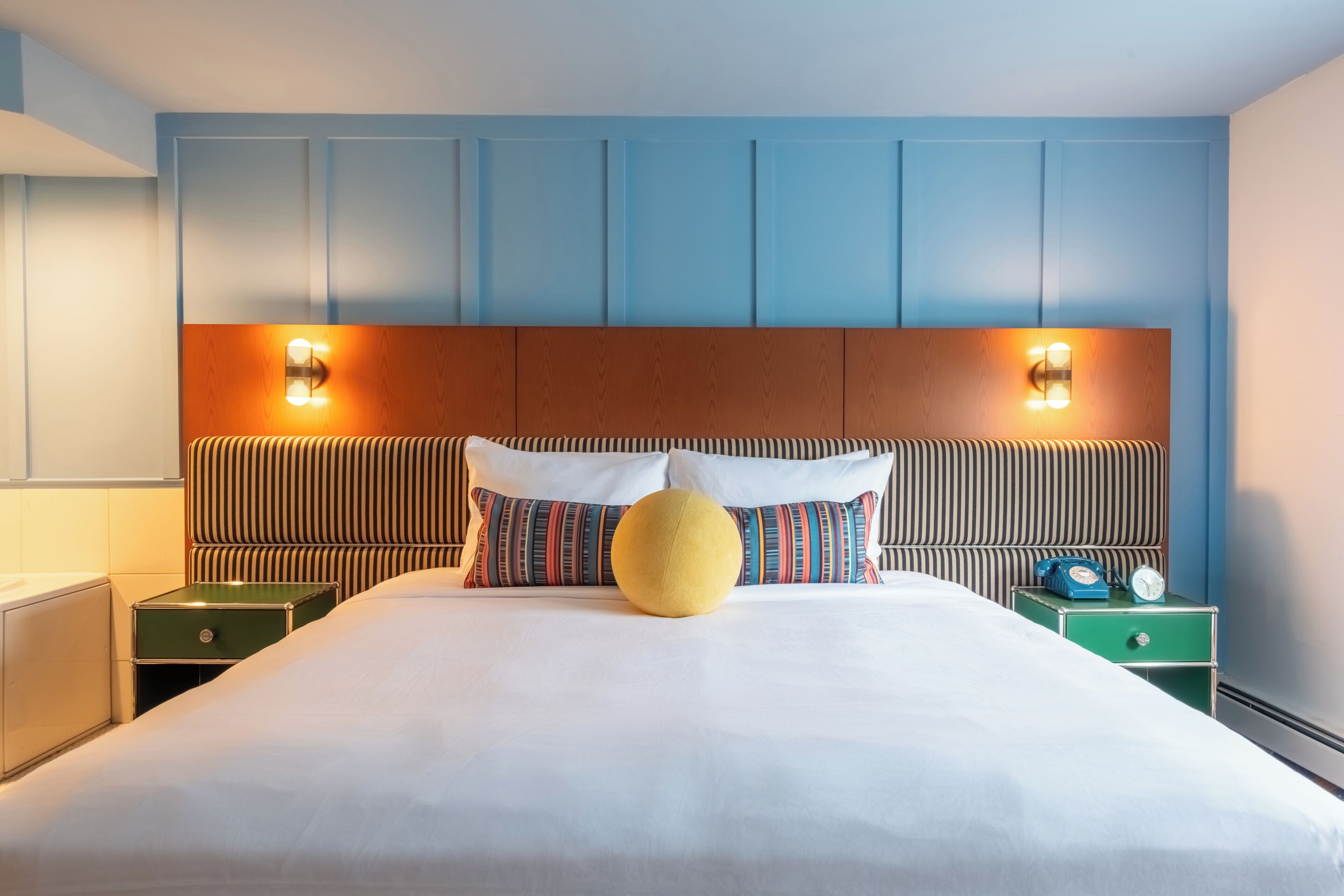 A hotel room with a large bed, striped pillows, a round yellow accent pillow, two bedside tables with clocks, and a wooden headboard with wall-mounted lights.