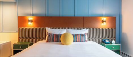 Hotelkamer met een groot bed, gestreepte kussens, een rond geel sierkussen, twee nachtkastjes met klokken, en een houten hoofdbord met wandlampen.