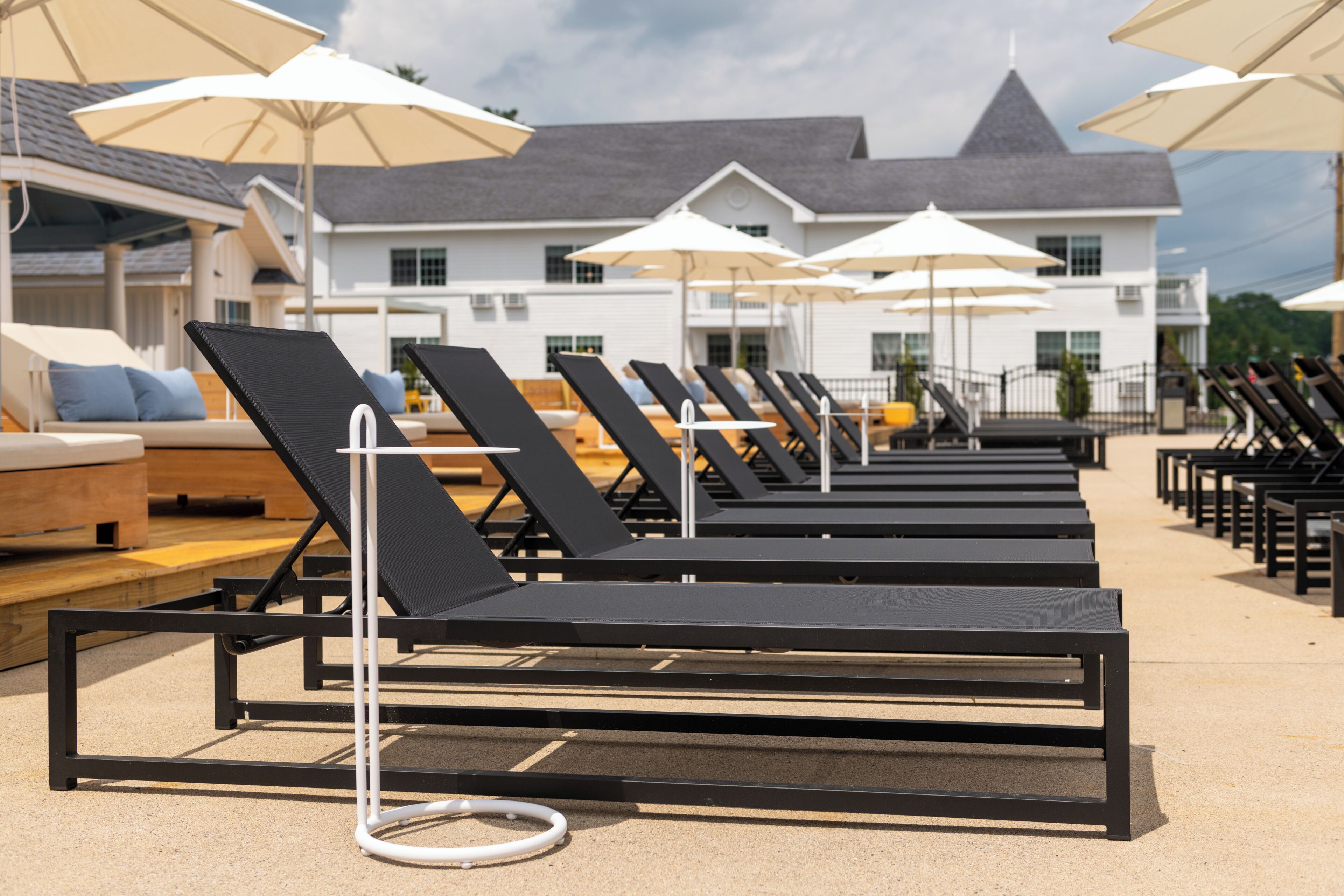 A row of sun loungers with umbrellas, a poolside area, and a building with a grey roof in the background.